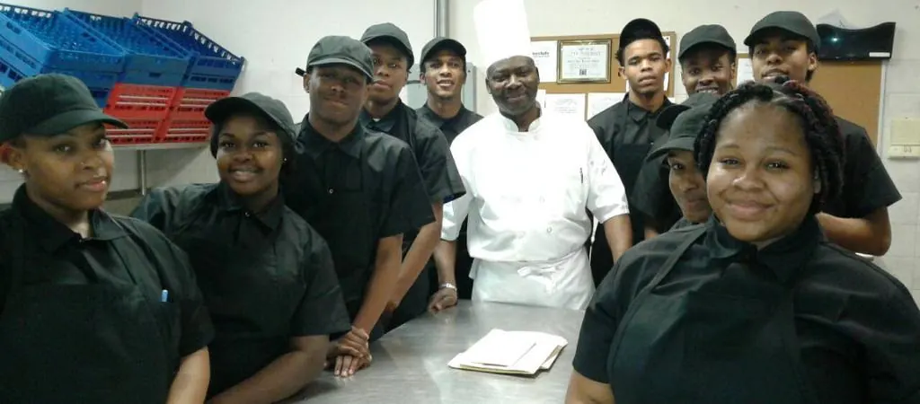 Mise En Place trainees in black uniforms gathered around a chef in whites